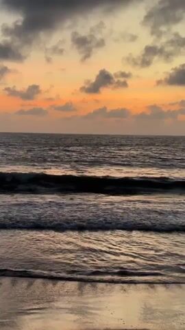 The video shows a sunset over the ocean. The sky is filled with clouds that are lit up with orange and yellow hues. The ocean is dark and wavy, and the waves are breaking on a sandy beach. The video is filmed from a low angle, making the ocean and sky appear vast and expansive. The sound of the waves crashing on the shore can be heard in the background.