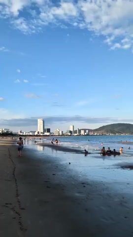 A vertical, eye-level shot captures a beach scene under a blue sky with scattered clouds. The foreground is a wet sand beach with reflections of the sky and distant buildings. A few people are visible walking on the sand and playing in the shallow water. In the mid-ground, there are more people swimming and wading in the ocean. A line of buildings, including a tall skyscraper, is visible on the horizon, with a green mountain in the background. The overall impression is of a sunny day at a populated beach.