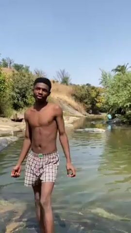 A young, slender man with dark skin, hair, and eyes stands in a shallow stream or river on a sunny day. He is shirtless and wears plaid boxer shorts. He looks into the camera and smiles at first, then looks down and to the side. The water appears calm, with a hint of green or brown, and reflects some sunlight. Behind the man are trees and a small hill. Another person is visible in the background, partially obscured by a rock. The sky is clear blue.