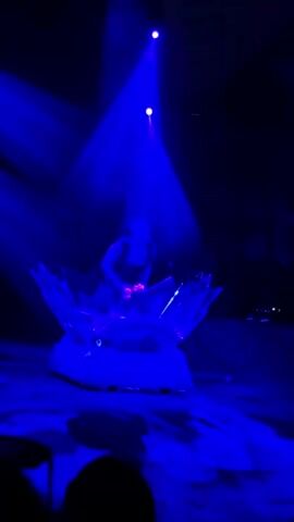 A performer stands on a raised platform surrounded by sharp, jagged, translucent blue props that resemble shards of ice. The performer is wearing a dark, sleeveless top and dark pants. They are juggling several small, purple objects. Two spotlights shine down on the performer from above, casting blue light onto the stage and the surrounding props. The stage floor is covered in a red carpet. In the foreground, the silhouettes of audience members can be seen. The performer moves their arms and body in a fluid motion as they continue to juggle. The lighting and props create a dramatic and visually striking atmosphere. The scene appears to be from a performance, possibly a circus or a theatrical show.