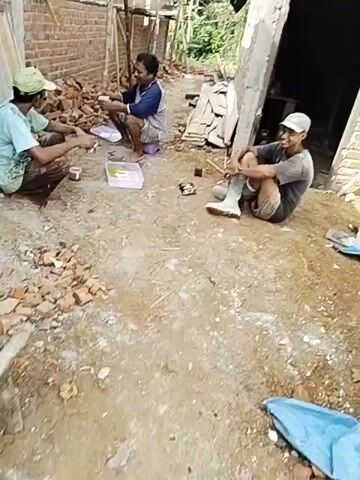 The video shows three men at what appears to be a construction site. The environment is outdoors with rough terrain and piles of bricks. Two of the men are seated on the ground, while one is squatting on a small stool. They are positioned in a way that suggests they are taking a break.

In the foreground, a man wearing a dark t-shirt, dark shorts, and tall white boots is sitting on the ground with his legs crossed. He is holding a cigarette in his right hand and appears to be talking or smiling. Next to him, on the ground, are a pair of white boots, a small package or box, and a cigarette butt. Further to his right, a wooden handle, possibly of a tool, is visible.

To the left of the foreground man, another man is squatting on a purple stool. He is wearing a blue and white long-sleeved shirt and light-colored shorts. He is holding something in his right hand and looking towards the man to his left. In front of him, on a white plastic tray, are several green circular items, possibly food or snacks, and a small glass of dark liquid.

A third man is visible further back, to the left of the man on the stool. He is wearing a light blue t-shirt with a design on it, dark pants, and a light green cap. He is sitting on the ground, facing the other two men, and appears to be engaged in conversation with them. He is holding a small object in his hands.

The background shows unfinished brick walls, wooden planks, and some foliage, indicating ongoing construction work. The lighting suggests it is daytime. The overall impression is a casual break among workers during a construction project.