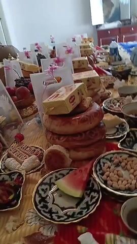A table is laden with various food items and gift bags. The food includes bread, pastries, fruits such as grapes, peaches, and watermelon, and nuts. The gift bags are white with pink ribbons. Several boxes of cookies and other packaged goods are also visible. A person is partially visible in the background, seated. The room appears to be indoors, with natural light coming from a window.