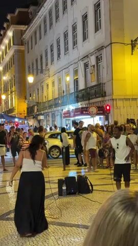 At night, on a cobblestone street lined with buildings, a group of people are gathered. A woman in a black skirt and white top stands on the left, facing away from the camera, with her arms outstretched. A man in a white tank top and black shorts stands on the right, holding a microphone and gesturing with his hands. He appears to be singing or speaking to the crowd. Behind them, a diverse group of men and women are standing and watching the performance. Some are holding up their phones to record. There are streetlights illuminating the scene, and some shops are visible in the background. The atmosphere seems lively and festive.