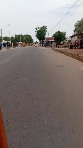 A video shows a street scene. The camera is mounted on a moving vehicle, likely a tricycle, facing forward. The street is paved and appears to be in a town or city. Several yellow tricycles, a common mode of transportation in some regions, are visible on the road, some moving in the same direction as the camera and others approaching. On the sides of the road, there are buildings and stalls that appear to be shops or market stands. Some people are standing near these stalls. The sky is overcast, suggesting a cloudy day. Trees are visible along the roadside. The overall impression is of a bustling, everyday street scene in a developing area.