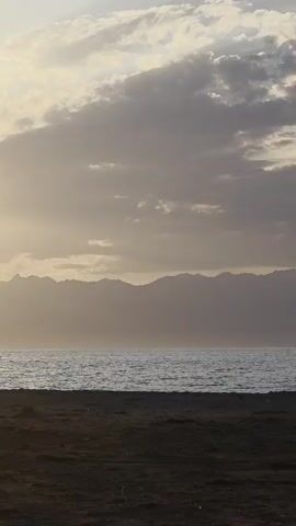 The video shows a beach scene at sunset. The sun is low in the sky, casting a warm glow over the water and clouds. Silhouettes of people are visible on the beach, some walking, some sitting, and some interacting with each other. The water is calm, and mountains are visible in the distance. The overall mood of the video is peaceful and serene.