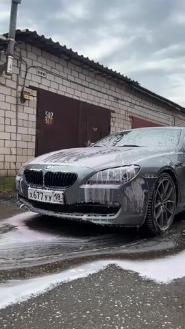 This media file shows a close-up, low-angle view of a grey BMW 6 Series coupe being washed. The car is covered in thick white and pink foam, with the foam dripping down the sides and pooling on the asphalt ground in front of it. The car's front grille, headlights, and license plate are clearly visible. The license plate reads "X 677 YY 18" and has "RUS" below it, indicating its Russian origin.

The car is parked in front of a brick garage with two large, dark brown doors. Above the doors, the number "502" is stenciled in white. Electrical conduits and junction boxes are attached to the side of the building to the left of the car.

The weather appears to be overcast with a cloudy sky. The overall scene suggests an outdoor car wash, likely at a private residence or a small car wash facility, given the utilitarian appearance of the garage. The foamy, sudsy water indicates the washing process is underway, creating a dynamic and active visual. There are no people visible in the frame. The time of day is not explicitly indicated, but the cloudy sky suggests it could be daytime.