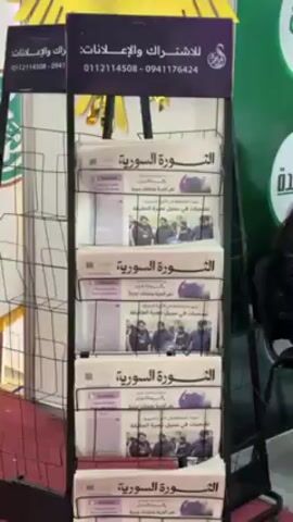The video shows a rack of newspapers in a convention hall with a red carpet. The newspapers are stacked in five rows and are titled "الثورة السورية". There are phone numbers for subscriptions and advertisements above the newspapers. In the background, people are walking around the hall, which appears to be part of a trade show or conference. Some booths are visible in the distance. A woman is sitting next to the newspaper rack.
