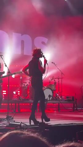 A female singer with short blonde hair, wearing a black vest, sparkly shorts, black tights, and platform heels, stands center stage on a platform, holding a microphone and singing. She is illuminated by red stage lights and thick white smoke. To her left, a male drummer is visible behind a drum set. To her right, a male guitarist is partially visible. The stage is equipped with various equipment, including microphones, speakers, and lighting rigs. The word "IONS" is visible in large white letters on a screen in the background. The overall scene depicts a live musical performance.