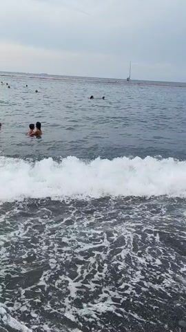 A video shows a view of a beach with people swimming in the ocean. The water is choppy with white waves crashing on the dark sand shore. In the background, a sailboat is visible on the horizon, along with some orange buoys. The sky is overcast.