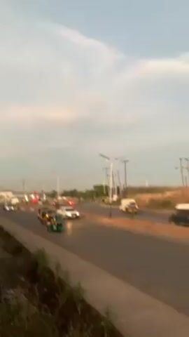 The video shows a street scene in what appears to be a developing country. The camera is positioned on a raised area, possibly an embankment or a low bridge, overlooking a busy road. The sky above is partly cloudy, suggesting either early morning or late afternoon light. Along the road, there are several vehicles, including cars, vans, and the distinctive three-wheeled auto rickshaws (also known as tuk-tuks). The vehicles are moving in both directions, indicating active traffic flow. Street lights are visible along the roadside, suggesting a partially urbanized area. The presence of power lines and poles further indicates infrastructure development. The surrounding environment includes some vegetation, suggesting that the area is not entirely urbanized and retains some natural elements. The general atmosphere suggests a busy, developing area with a mix of urban and semi-rural elements.