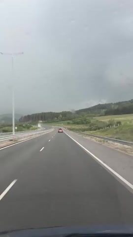 The video shows a first-person perspective from a vehicle driving on a highway on a cloudy day, likely near Dragoman, Bulgaria. The road appears wet, suggesting recent rain or drizzle. A red car is visible ahead in the same lane. The highway curves gently through a landscape of rolling hills covered with green fields and dense forests, mostly coniferous trees. Guardrails line both sides of the highway. The sky is overcast and grey. No people are visible, and no readable text can be discerned.
