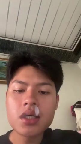 A close-up shot shows a young man with black hair, looking directly at the camera. He is wearing a black t-shirt. In the background, a white ceiling with horizontal lines is visible, along with a framed picture on the left and some white fabric on the right. The man brings a cigarette to his lips and exhales a ring of smoke, which dissipates in front of his face.