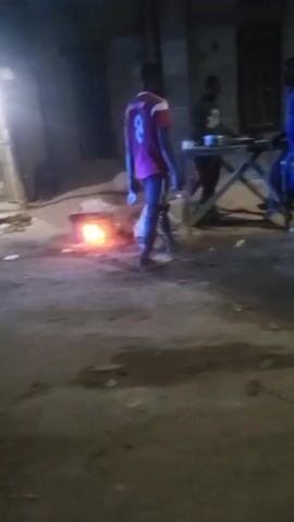 The video shows an outdoor area at night with several people present. In the foreground, there is a small fire burning on the ground. Three individuals are gathered around a table further back. One of them is wearing a red jersey with the number 8 on the back. The area seems to be somewhat dimly lit with what appears to be debris or litter on the ground. A person is seen riding a motorcycle in the background.