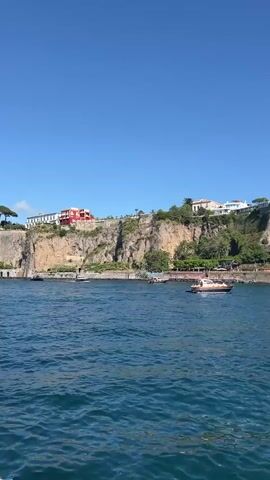 The video pans across a body of blue water, showing a cliffside town with buildings and lush greenery. Several boats are visible in the water, including a large white yacht and a smaller speed boat. The sky is clear and blue. The scene appears to be a sunny day in a coastal location.