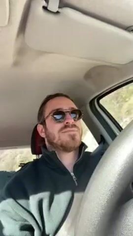 A man with a beard and sunglasses is driving a car. He has long hair pulled back. He wears a dark green fleece jacket. The car interior is visible. He looks ahead and to the side as he drives. A snippet of music is heard.