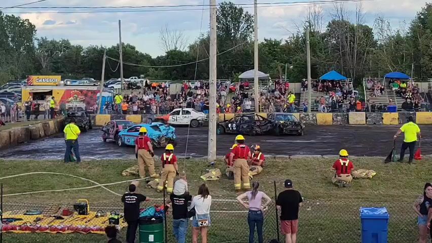 A demolition derby event is underway. In the foreground, several individuals in protective gear, likely firefighters or safety personnel, are positioned on a grassy area. They are dressed in yellow hard hats and red or black uniforms. Some are seated, others are standing. Equipment such as hoses and what appears to be a generator are visible nearby. Beyond this area is a dirt track, the derby arena, where several heavily damaged cars are located. Some cars are upright, while others are overturned or significantly crushed. In the background, spectators are seated in tiered bleachers, observing the event. A food vendor or concession stand is visible on the left side of the image. The sky is overcast with a bright, possibly late afternoon or early evening light. The overall atmosphere suggests an active event with safety precautions in place.