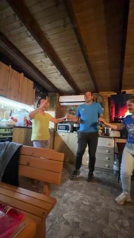 Three people are dancing inside a cabin. A woman in a yellow shirt and grey pants is in the foreground, facing away from the camera, with her arms outstretched as she dances. A man in a blue shirt and dark pants is to her left, holding hands with her and another woman who is wearing a blue t-shirt and jeans. Behind the dancers is a kitchen area with wooden cabinets and a microwave. To the right of the dancers is a TV showing a fireplace scene. A wooden bench is visible in the foreground. The cabin has a wooden ceiling.