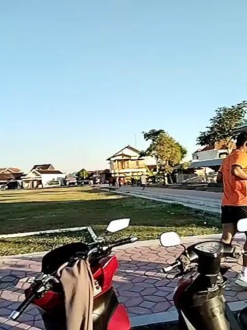 The video opens with a static shot of a paved area with several motorcycles parked. A tall pole runs vertically through the right side of the frame. In the background, there is a building and some trees. As the video progresses, the camera pans to the left, revealing a grassy field and more buildings. People are seen walking and running in the distance. The camera continues to pan and tilt upwards, showing a clear blue sky. A few more motorcycles and a bicycle are visible in the foreground. The scene appears to be an outdoor public space, possibly a park or a community area, with a clear day and bright sunlight. The overall atmosphere is calm and casual, with people engaged in various activities.
