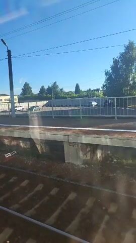 The video shows a view from a train window. The train is moving along a track. Outside the window, there is a train platform with a concrete edge and a gravel ballast. Beyond the platform, there is a white metal fence, a paved area, and a row of cars parked. Further in the background, there are trees and buildings. The sky is clear and blue. There are overhead power lines visible in the sky. The video is short and the camera is panning slightly to the right.