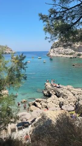 The video shows a scenic bay with clear turquoise water and rocky shorelines. Several people are visible enjoying the water and sun. Some are swimming, while others are lounging on rocks or beach chairs. A few small boats are anchored in the bay. Lush green trees frame the foreground of the shot. The overall scene depicts a popular tourist destination for swimming and relaxation.