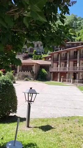 This is a panning shot of a hotel building and its surroundings in Setcases, Spain. The building is a multi-story structure with a stone facade and wooden balconies. It appears to be nestled in a mountainous area, with lush green trees and hills in the background. The scene is set during the day, with bright sunlight casting shadows on the paved courtyard. A lamppost stands in the foreground, and a well-maintained bush and grassy area are also visible. The overall impression is one of a peaceful, rustic retreat. There are no people visible in the shot. The time of day appears to be late morning or early afternoon, given the length and angle of the shadows. The weather seems to be clear and sunny. The architecture of the building, with its stone and wood elements, suggests a traditional mountain style, common in regions like the Pyrenees where Setcases is located. The vibrant green foliage indicates a time of year with ample vegetation, likely spring or summer. The image conveys a sense of tranquility and natural beauty.