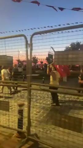 Here is a concise description of the video:

The video shows a crowd of people, possibly in a public space. A metal fence runs along the foreground of the scene.  Many individuals are walking, and at least one person is holding a Turkish flag. Decorative flags are strung overhead.  The time of day appears to be dusk or evening, and streetlights illuminate the area. Buildings are visible in the background.