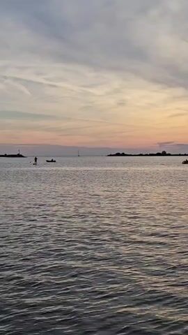 The video shows a serene body of water with a sky painted in soft pastel colors, indicating either sunrise or sunset. The water is rippled, suggesting a gentle breeze. In the distance, there are two small islands or landmasses, one on the left with a lighthouse-like structure and another on the right. Between these, on the water, a person is paddleboarding, with a paddle in their hand, appearing to be standing upright. To the right of the paddleboarder, there is a small boat with at least one person inside. Further out in the water, a sailboat is visible. The overall scene conveys a sense of tranquility and outdoor recreation.