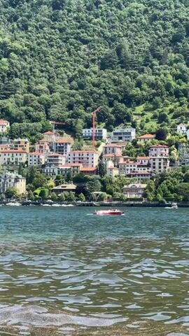 The video shows a town nestled on a steep, green hillside overlooking a body of water. The town consists of closely-packed buildings, some with red-tiled roofs, interspersed with greenery. Two red construction cranes are visible among the buildings. In the foreground, the water ripples slightly, and a small red and white boat crosses the scene. Other boats are docked near the shoreline. The hillside is densely covered with trees.