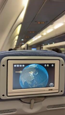The video shows the interior of an airplane cabin from the perspective of a passenger seat. Rows of blue cloth seats fill the frame, each with a personal entertainment screen displaying a map of the world. The map shows the route of the flight from Istanbul to Beijing. Other passengers are seated, but their faces are not visible. The cabin is lit by overhead lights.
