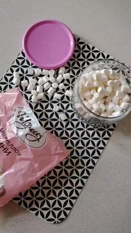 The video shows a table with a black and white patterned placemat on it. There is a pink lid on the left side of the placemat. To the right of the lid, there are many small white marshmallows scattered on the placemat. In the upper right corner of the placemat, there is a clear glass jar filled with mini marshmallows. In the lower half of the placemat, there is a partially opened pink bag of mini marshmallows. A hand is reaching into the bag, as if to take out marshmallows. The text on the bag is in Russian and says "Маршмеллоу Мини," which translates to "Mini Marshmallow." The overall scene suggests preparation for a snack or dessert involving marshmallows.