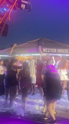 A group of people are dancing outdoors at night, with colorful lights illuminating the scene. A sign in the background reads "NESTER HOUSE BUNGALOW". Foam is visible on the ground, suggesting a foam party. The sky is dark with some orange lights visible in the distance. The people are dressed in casual summer clothing, and some appear to be children. They are moving their arms and bodies to the music, and some are holding their phones up, possibly recording the event. The overall atmosphere seems festive and energetic.