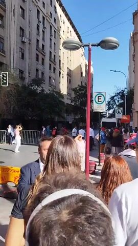 The video shows a group of people standing behind a barrier on a city street, observing something happening further down the street. The camera appears to be held by someone in the crowd. The group of people, seen from the back, are diverse, including men and women of different ages and hairstyles. Some are looking toward the end of the street where the focus of attention seems to be. The barrier is made of metal poles and horizontal bars. Beyond the barrier, there are more people, including some who seem to be in uniform. A white van is parked on the street. On the left is a tall building with many windows and air conditioning units visible. On the right, another building is visible. Street signs are attached to a red pole in the foreground. The sky is blue. The general impression is of a gathering of people watching an event or situation unfold on a city street.