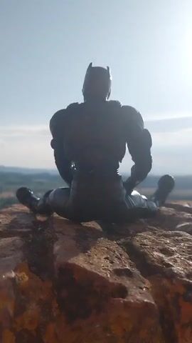 A Batman action figure is positioned in a seated split on a rocky outcrop. The figure faces away from the camera, looking out over a valley and distant hills. The sky is bright blue with a few wispy clouds, suggesting a sunny day. In the background, a village with red-tiled roofs is visible, nestled amongst green trees and fields. A colorful banner or flag, possibly with geometric patterns, is also visible in the mid-ground, fluttering in the breeze. The scene appears to be set in a rural European landscape. The time of day is likely midday or early afternoon, given the bright sunlight and clear sky. The overall mood is contemplative, with the iconic superhero figure appearing to be taking a moment of pause and reflection in a picturesque setting.