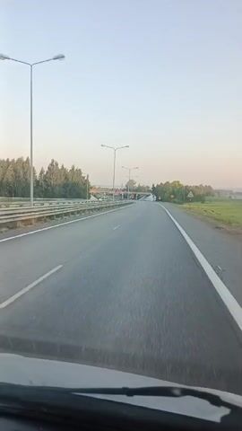 The video is a first-person perspective from inside a vehicle driving on a highway. The road has two lanes in the direction of travel, with a guardrail separating the lanes from the opposing traffic. The sky is clear and appears to be early morning or late afternoon. There are trees and fields on either side of the highway. Streetlights are visible along the road, and there are signs indicating a speed limit of 50. In the distance, an overpass can be seen. The overall atmosphere is calm and serene, suggesting a peaceful journey.