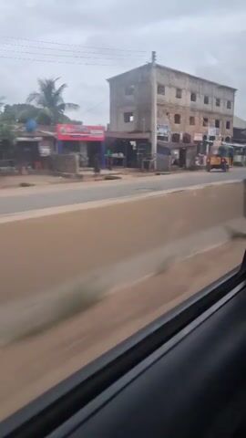 The video shows a view from inside a vehicle, looking out at a road with buildings and various vehicles. The scene is somewhat blurry due to motion.  In the foreground, a beige barrier runs horizontally across the frame.  On the other side of the barrier, a paved road is visible.  Several buildings line the road, varying in style and condition, some appearing to be shops or businesses.  Traffic includes a yellow auto-rickshaw, a white van, motorcycles, and a red truck with a blue trailer.  People are visible near the shops.  The sky is overcast. The overall impression is of a busy, possibly developing, urban or semi-urban environment.