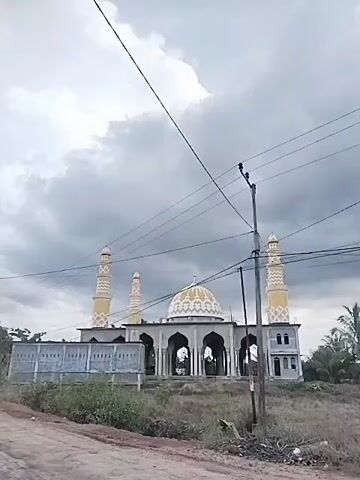 The video shows a mosque under construction on a cloudy day. The mosque has a central dome and four minarets. The dome is decorated with a geometric pattern. The mosque is surrounded by a partially constructed wall and some vegetation. There is a dirt road in front of the mosque. Power lines run across the front of the scene. The sky is overcast. There is an echoing noise during the footage.