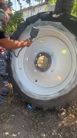 A person wearing a black t-shirt, shorts, and a protective mask over their face is spray painting a large white tractor wheel. The person is holding a spray gun connected to an air compressor hose, applying white paint to the wheel. The wheel is large and black, with a white rim that is being painted. The background shows an outdoor environment with trees and dirt. The person is focused on the task, carefully spraying the paint.