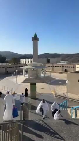 The video depicts an outdoor view of a mosque, specifically the Masjid al-Bay'ah located in Mina, Saudi Arabia. Several people dressed in white ihram clothing are walking down a set of stairs towards the mosque courtyard. The mosque has a tall, cylindrical minaret with a green dome on top. There's also a pavilion-like structure in the courtyard, with people milling around it. The overall environment is bright and sunny, with clear blue skies. The architecture of the surrounding buildings is simple and functional. The people are likely pilgrims undertaking religious rituals. The video provides a glimpse into the atmosphere of a significant Islamic site during a pilgrimage season.