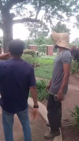 The media file shows two men standing in a garden in Iju, Nigeria. One man, on the left, is wearing a dark blue t-shirt and blue jeans, with a dark baseball cap. He is facing away from the camera, looking towards the other man. The second man, on the right, is wearing a grey t-shirt, dark trousers, and brown boots. He is wearing a large, wide-brimmed straw hat that obscures his face. He is holding a mobile phone in his right hand and appears to be speaking or laughing. The setting is a lush garden with various plants, including large leafy plants in the foreground and bushes and trees in the background. A red brick pathway is visible behind the plants. In the distance, a fence and some buildings can be seen. The lighting suggests it is daytime, possibly late afternoon, with soft sunlight filtering through the trees. The overall mood appears relaxed and casual, with the man in the straw hat displaying an expression of amusement. There is no visible text in the media file.