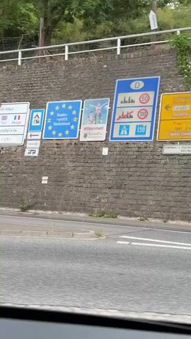The video begins with a close-up shot of a brick wall adorned with various road signs. On the left, a sign with stars signifies European Union membership. Next to it, a sign displays "Willkommen!" (Welcome!) and the state of Rhineland-Palatinate, Germany. A prominent blue and white sign with a German flag symbol indicates speed limits: 50 km/h in built-up areas, 100 km/h outside built-up areas, and 130 km/h on autobahns. A yellow sign points towards Trier and Langsur, with further details for Trier and Saarlouis. The camera then pans to the right, showing a smaller sign with place names. The view shifts to the interior of a car, showing the dashboard and windshield. The car is driving on a road next to a similar brick wall, passing under a bridge. The landscape visible through the windshield reveals a hilly area with houses and trees. The car continues driving on a curving road, passing other vehicles and a small bus stop shelter. The scene appears to be in a European country, likely Germany, given the road signs.