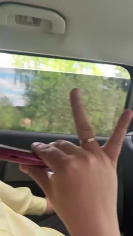 Inside a car, a person wearing a yellow shirt and a ring on their finger exhales a large plume of vapor from a red vape pen, which they then use again. The car window shows a view of green trees and a slightly cloudy sky. The interior of the car is dark.