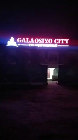 The video shows a building at night with a brightly lit sign that reads "GALA OSIYO CITY" in red lettering. Below this, smaller white letters read "TURAR JOY MAJMUA." To the left of the text is a stylized white silhouette of a city skyline. The building itself is dark and mostly obscured, but a doorway is visible with light emanating from within. The ground in front of the building appears to be unpaved.