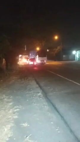 At night, on a two-lane road, a motorcycle with a rider wearing a helmet and a light-colored jacket is traveling away from the camera. The motorcycle has a red taillight and its brake lights are illuminated. The motorcycle passes by a parked truck. The truck is white with a blue stripe on its side and its headlights are on. The truck appears to be parked on the side of the road. There are trees and foliage on the left side of the road and a building on the right side of the road. There is a street light illuminating the scene. The road surface is asphalt.