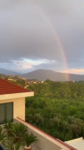 The video captures a panoramic view of a scenic landscape, likely from a high vantage point such as a balcony or terrace in Trebesin, Montenegro. In the foreground, the corner of a building with a distinctive red-tiled roof, light-colored walls, and green window shutters is visible on the left. Potted palm-like plants adorn a balcony railing in the lower left.

The main focus of the scene is a vibrant rainbow arcing across a partly cloudy sky. The sky is predominantly grey with darker clouds, indicating recent rain, but patches of lighter sky and soft, warm light suggest late afternoon or early evening.

The mid-ground and background are dominated by rolling hills densely covered in lush green trees and vegetation. Numerous white or light-colored houses with red roofs are scattered across these hills, indicating a residential area. In the distance, a body of water, likely the Bay of Kotor or the Adriatic Sea, can be seen, partially obscured by the hills. Beyond the water, more mountains or hills rise against the horizon.

The camera performs a slow, subtle pan to the right throughout the video, gradually revealing more of the landscape and the full arc of the rainbow, which extends from the left side of the frame towards the right, appearing to touch down in the distant green hills. The scene captures the serene aftermath of a rain shower, highlighted by the striking natural phenomenon of the rainbow. No visible text is present in the video.