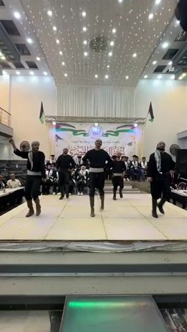 A group of five men are performing a dance on a stage. They are all wearing black shirts with white sashes around their waists and dark pants tucked into boots. They also have white head coverings. The men are in sync as they move their arms and legs. In the background, there is a banner with Arabic writing and images of Palestinian flags. Several people are seated in the background, some wearing graduation caps and gowns. The stage is lit with bright lights, and there are string lights hanging from the ceiling. The overall environment appears to be an indoor venue, possibly a hall or auditorium.