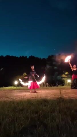 Two women are performing with fire in a grassy outdoor area at night. The woman on the left is wearing a black and red dress and is twirling two flaming props, creating circles of fire. The woman on the right, also in a black and red outfit, is holding a single flaming prop above her head. In the background, there is a body of water, trees, and lights, suggesting a park or waterfront location. A portable speaker is visible to the left of the stage. The sky is dark blue with a faint haze.
