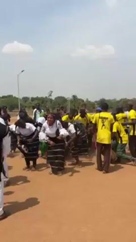 The video shows a group of people gathered in an open outdoor space. The ground appears to be reddish dirt. There are trees and vegetation in the background, along with a few structures, including a light pole. The sky is mostly clear. The group consists of men and women. Some of the women are wearing matching outfits with white tops and skirts with black and white horizontal stripes. Many of the men are wearing yellow t-shirts with a dark graphic on the back. Some men and women are wearing other traditional clothing. The group is gathered around a percussionist and appears to be dancing and moving to the beat of the drums. The camera moves to capture different angles of the group and the dancing.