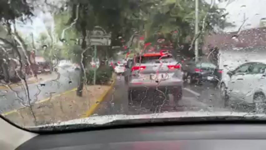 The scene is captured from inside a vehicle, looking out through the windshield. It is raining heavily, with raindrops visibly streaming down the glass. The windshield wipers are in motion, clearing the water. Through the blurred view, several cars can be seen ahead on the road, with the car directly in front being a silver SUV. The street is wet and glistening, with a yellow-painted curb separating the road from a green, tree-lined area. There are street signs visible in the periphery, though the text is difficult to read due to the rain and distance. The weather appears overcast and visibility is reduced due to the heavy rain.