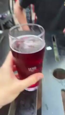 Here is a description of the video.

The video shows a person holding a glass of red liquid. The camera turns upward to show the face of a fair-skinned person with short brown hair and green eyes. A fair-skinned woman with blonde hair is seen in the background. The person in the foreground is drinking from the glass. The video then shows a close-up of a red, lattice-like object on a table.