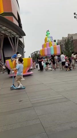 The video shows an outdoor plaza with people milling about. In the foreground on the left is a large, brightly colored inflatable tea cup with a person sitting on its rim. Behind the tea cup, a larger, tiered inflatable structure resembling a cake is visible, displaying the word "DREAM" with colorful letters and a green ribbon. Several people are gathered around this structure, including adults and children. The background features tall apartment buildings and trees under an overcast sky. People are walking, standing, and interacting with each other. One person in the middle ground is carrying a baby in a stroller. To the right, a man in a black shirt and pants is walking towards the camera while looking at his phone. Other individuals are scattered throughout the plaza.